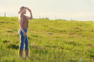 A woman wearing a cowboy hat stands in a sunlit countryside field.
