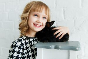 A smiling young girl with blond hair cuddles a cute black rabbit indoors.