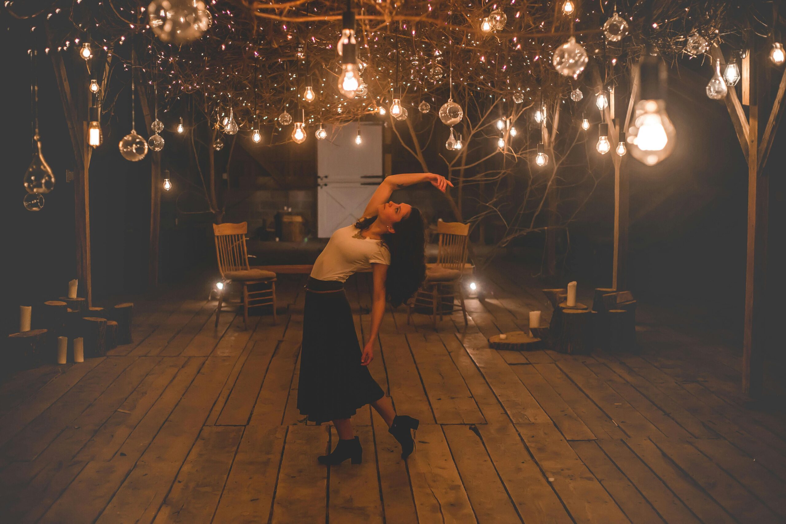 A woman gracefully dances under warm, illuminated bulbs in a rustic wooden interior.