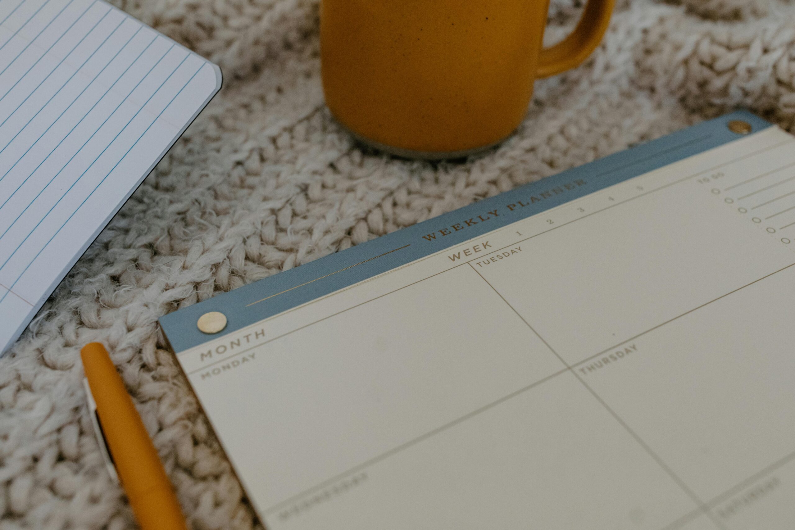 A cozy workspace featuring a weekly planner, pen, and coffee mug on knitted fabric. Perfect for planning.