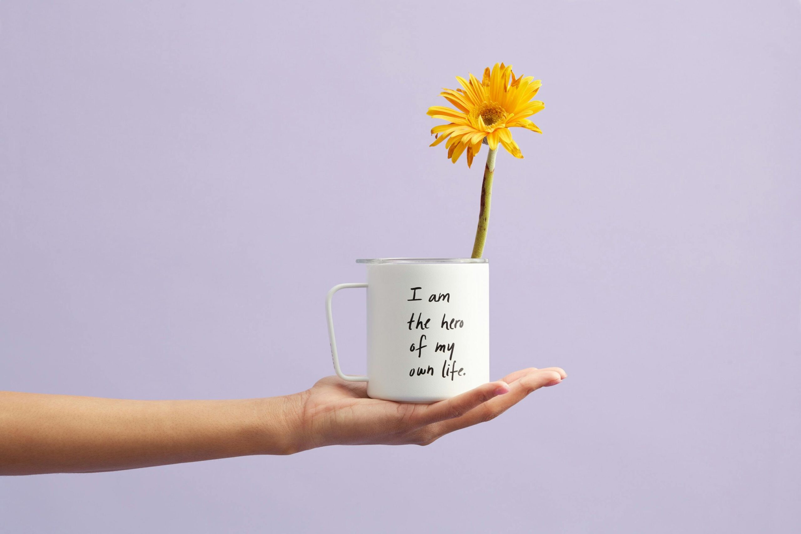 Hand holding a mug with a sunflower and motivational quote, set against a purple background.